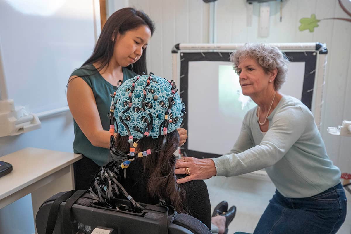 A woman sits with her back to the camera wearing a helmet covered with electronics. Two more women stand either side